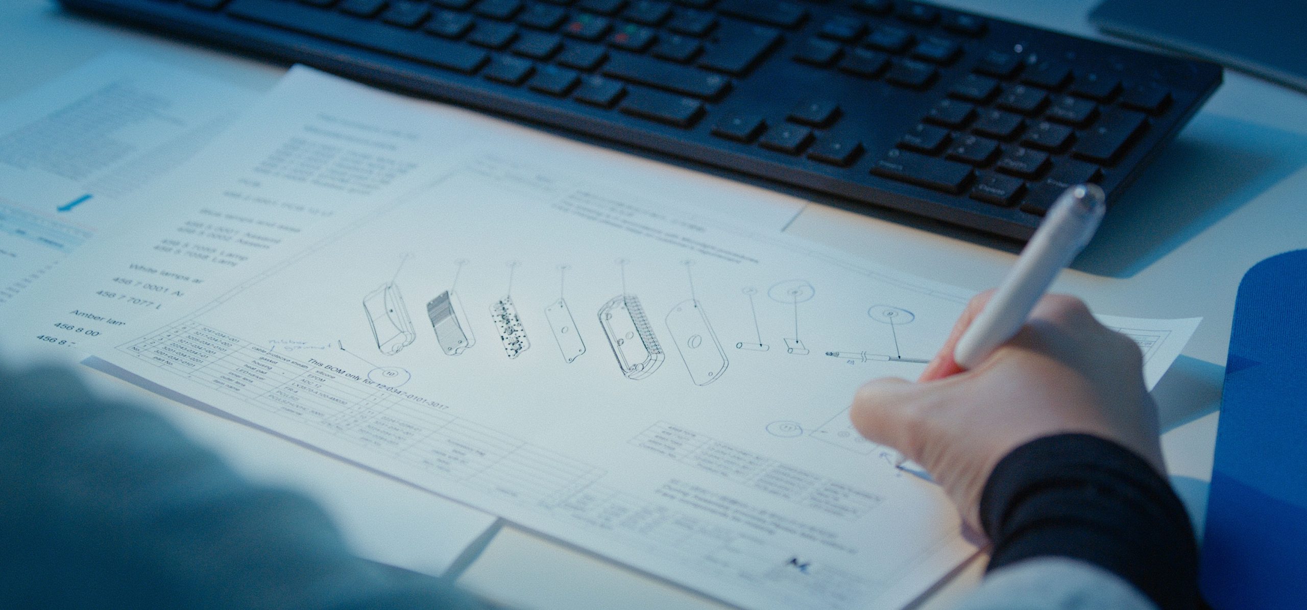 Hero Image Paper Schematics Close Up on Table with Computer Keyboard. Hand Holding Pen Writing Onto the Sheet