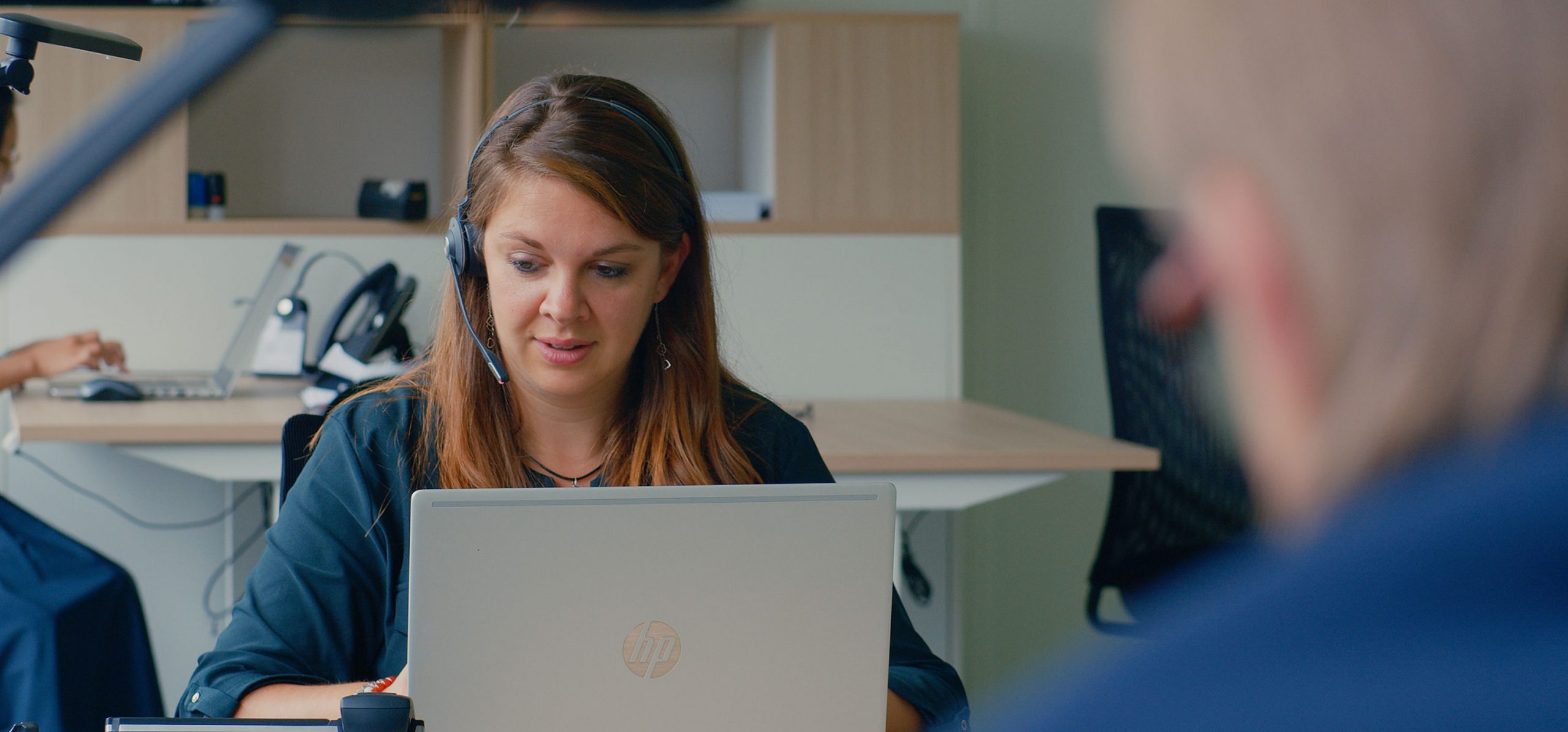 Hero Image Person wearing headset is taking a phonecall while looking down at a laptop screen, over the shoulder shot of an out of focus figure in foreground