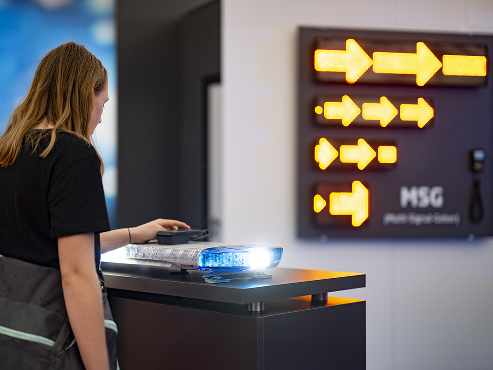 Over the shoulder shot of a person interacting with a blue LED lightbar, the background is out of focus and shows a display of amber scrolling LED message signs.