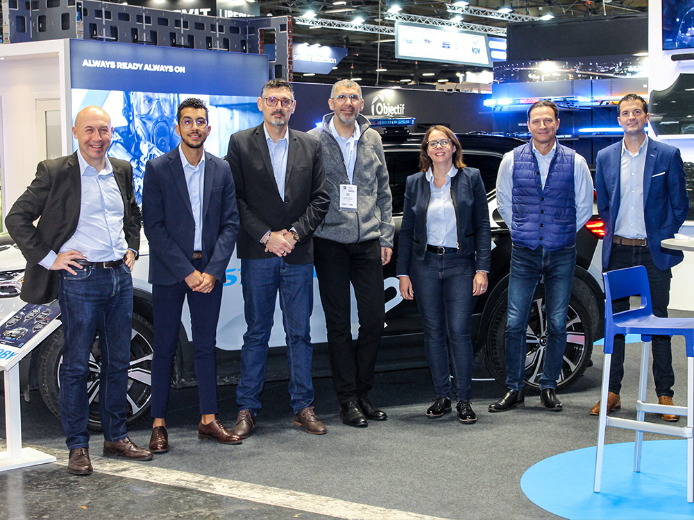 Group photo of seven Standby Group employees on a Standby Mercura exhibition stand, facing the camera and smiling in front of a Police car.