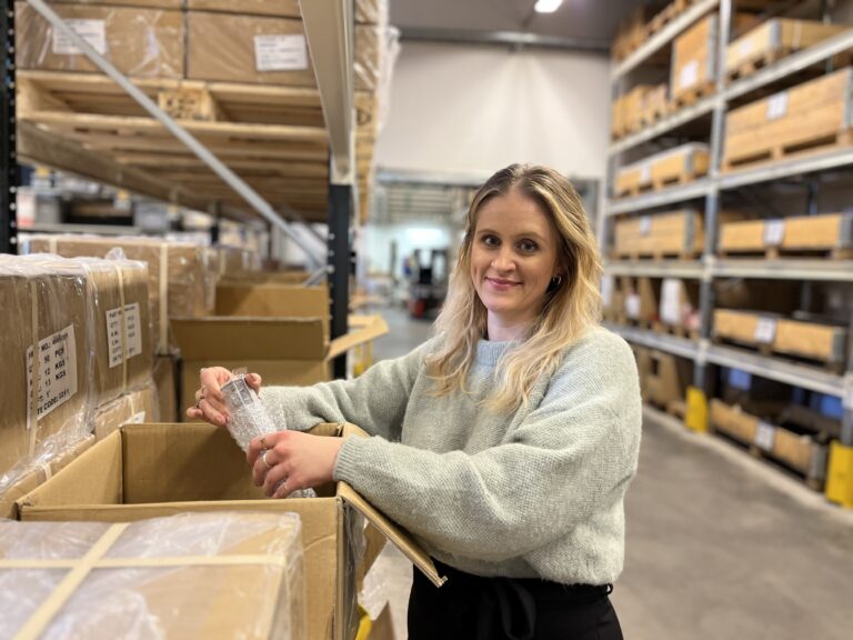 En medarbetare står på ett lager och håller i en produkt som hon tagit upp ur en låda.