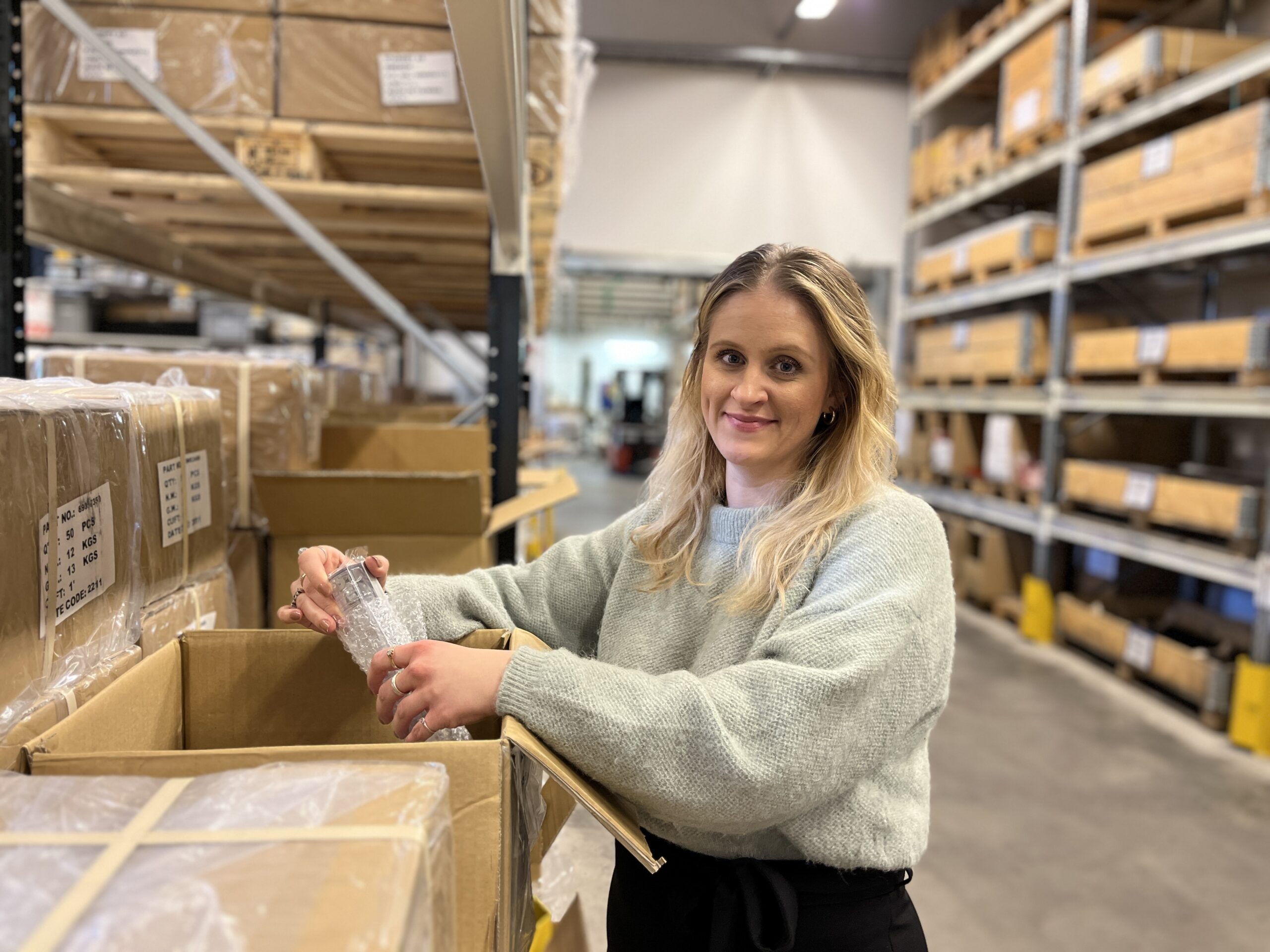En medarbetare står på ett lager och håller i en produkt som hon tagit upp ur en låda.
