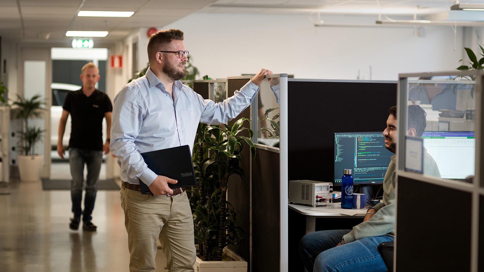 En säljare står vid en kollegas skrivbord och pratar medan en annan kollega kommer gående längre bak i bilden.